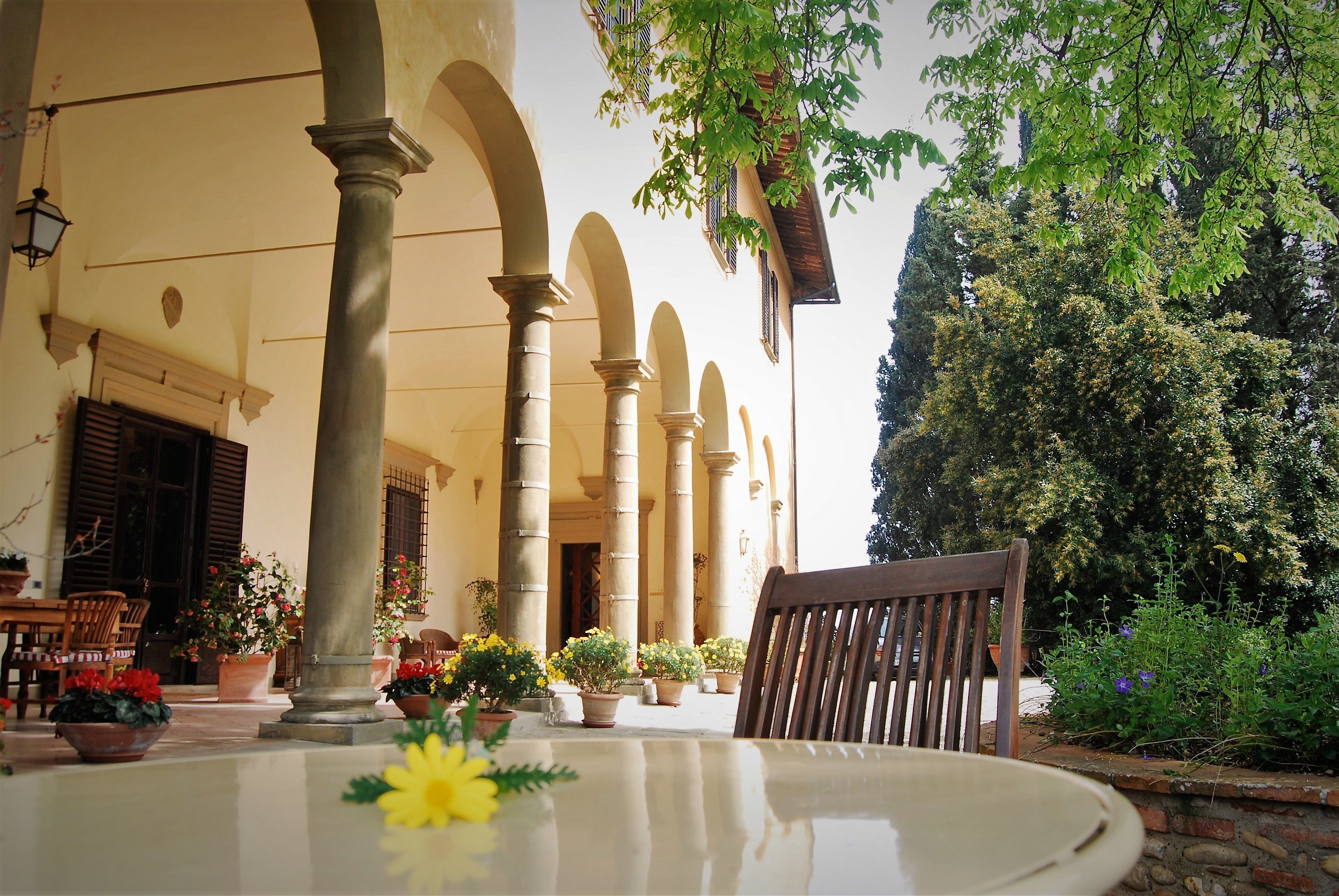 A covered outdoor seating area with a table and chairs, surrounded by columns and potted plants.