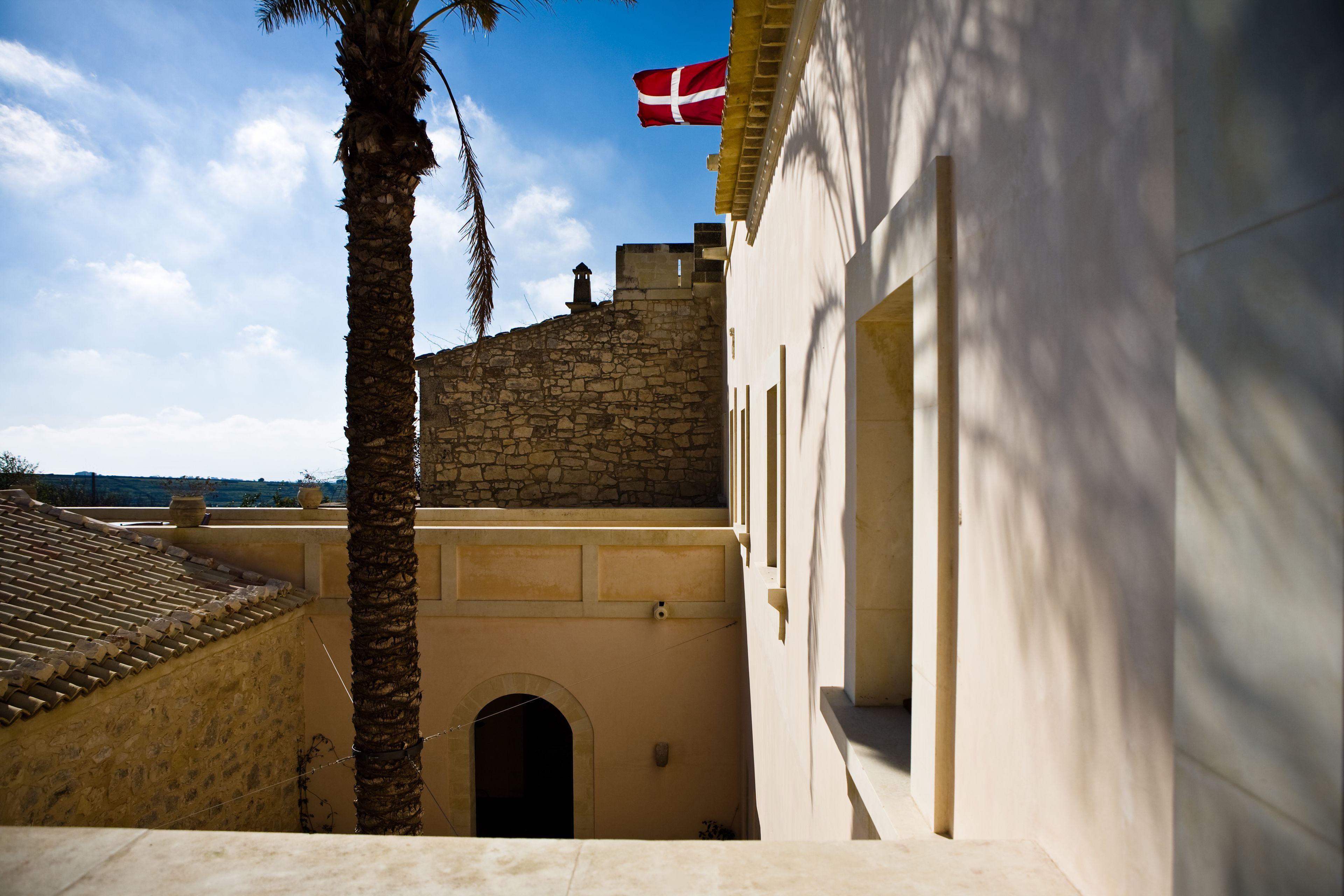 A narrow alleyway with a stone wall, a palm tree, and a building with a flag.