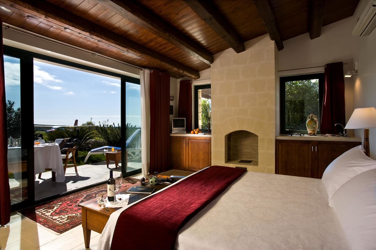 A bedroom with a large bed, a wooden ceiling, a fireplace, and a view of the outdoors.