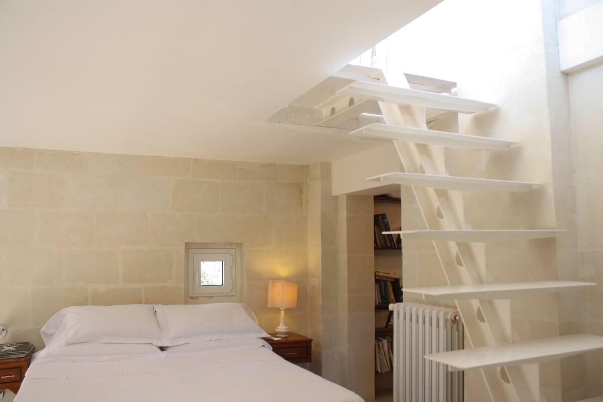 A bedroom with a bed, a nightstand, a lamp, a radiator, and a staircase.