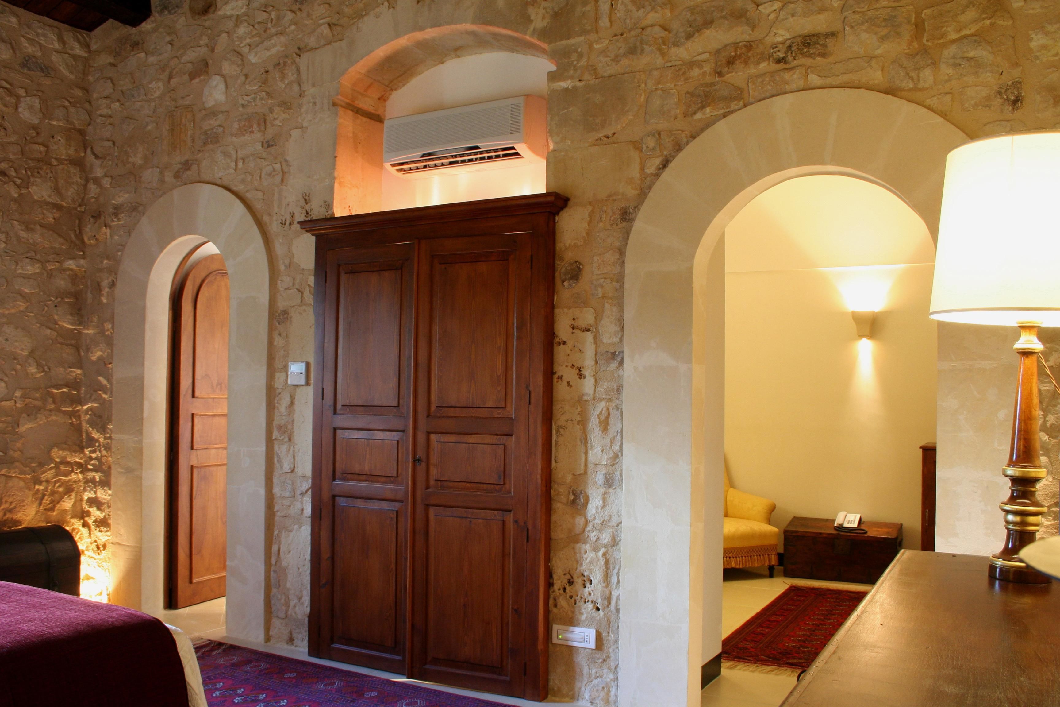 A room with stone walls, a wooden wardrobe, a bed with a red blanket, and a lamp.
