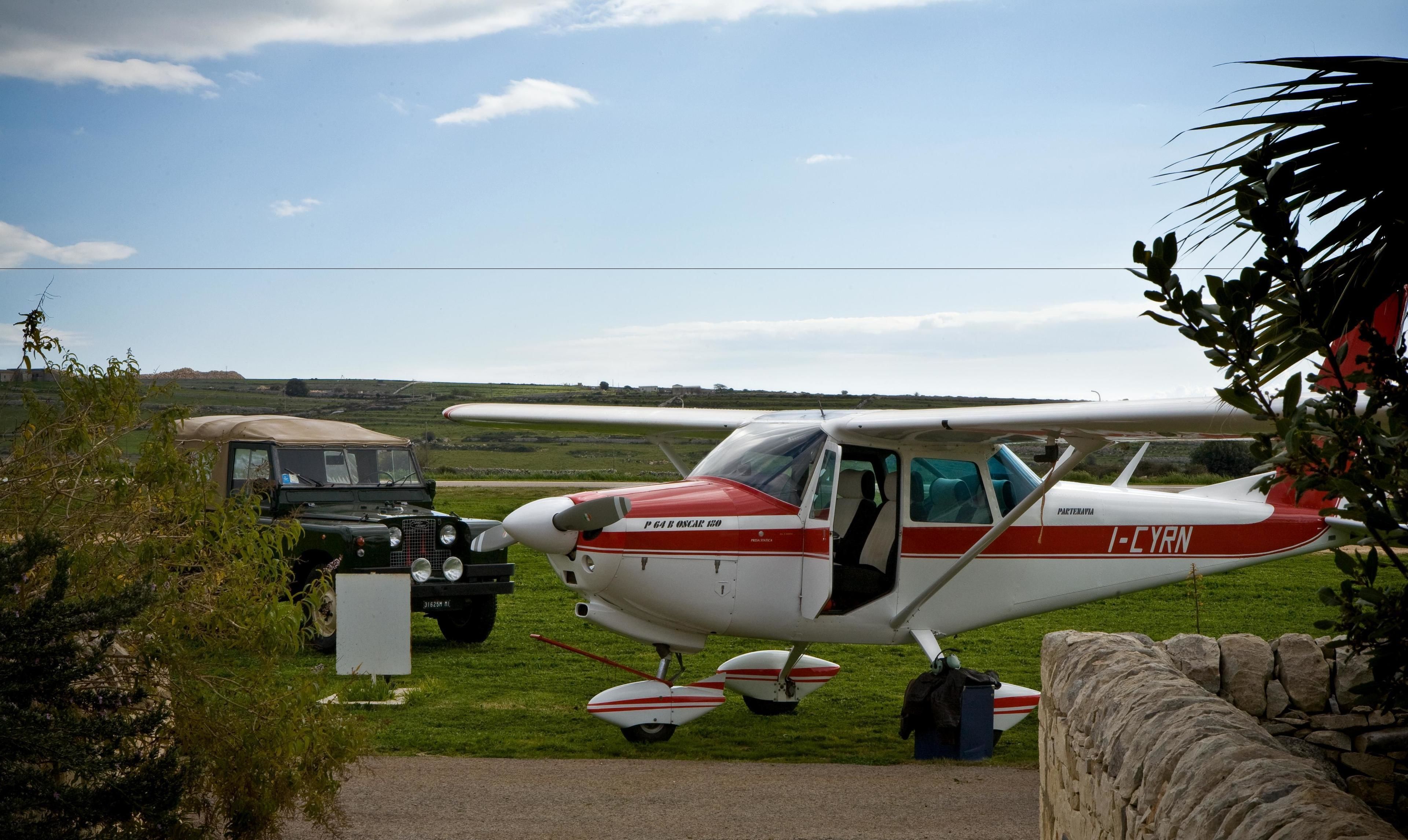 A small red and white aircraft parked on a grassy field with a vintage car nearby.