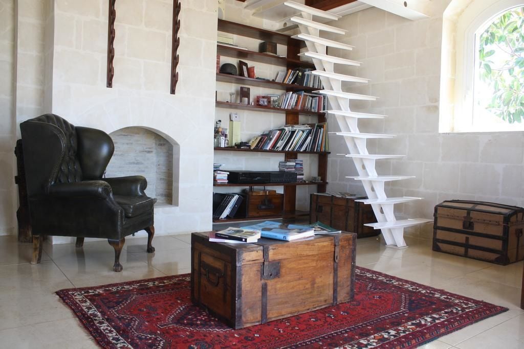 A cozy living room with a fireplace, a black leather armchair, a wooden coffee table with books, a white ladder bookshelf, and a red patterned rug.