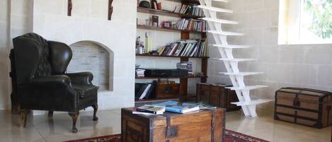A cozy living room with a fireplace, a black leather armchair, a wooden coffee table with books, a white ladder bookshelf, and a red patterned rug.