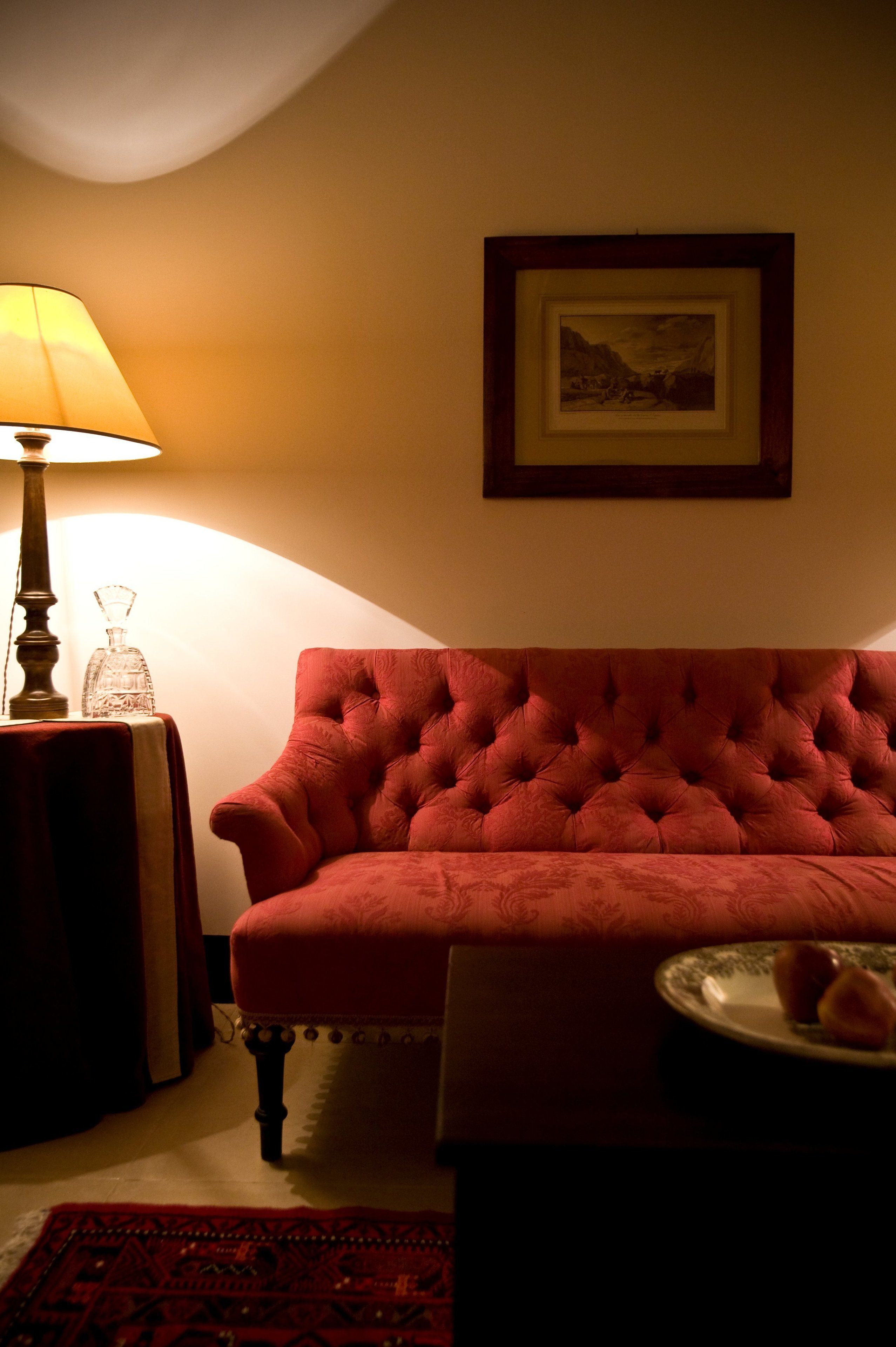 Une pièce avec un canapé rouge à motifs capitonnés, une petite table avec une lampe, un tableau encadré au mur et une assiette de nourriture sur une table.