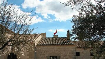 Un edificio de piedra con techo de tejas, una bandera y un patio empedrado con plantas en macetas.