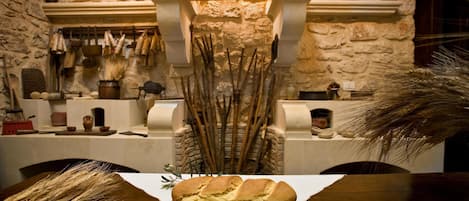 Cocina rústica con paredes de piedra, una mesa de madera y una hogaza de pan sobre un mantel blanco.