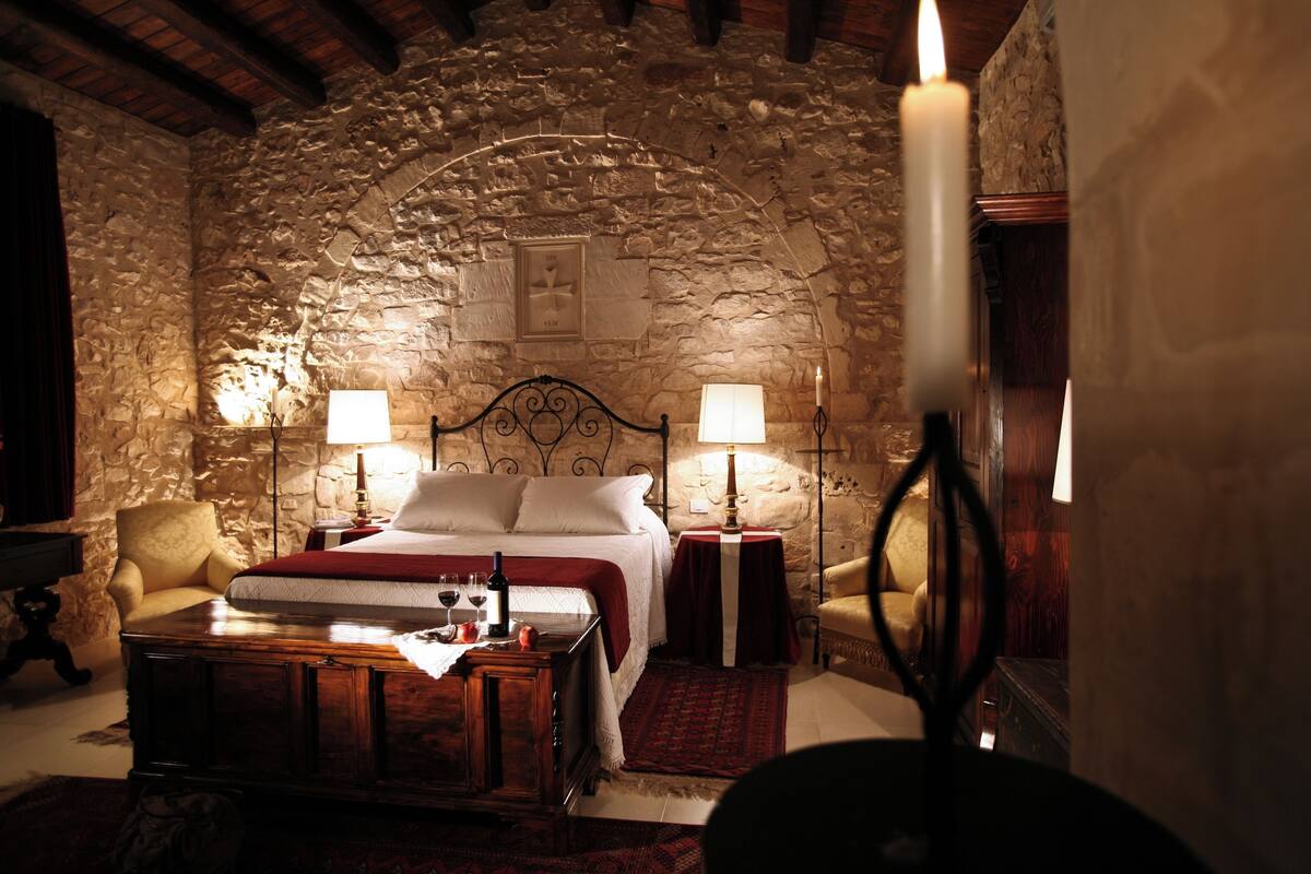 A stone-walled bedroom with a bed, two bedside tables, two lamps, and a wooden nightstand.
