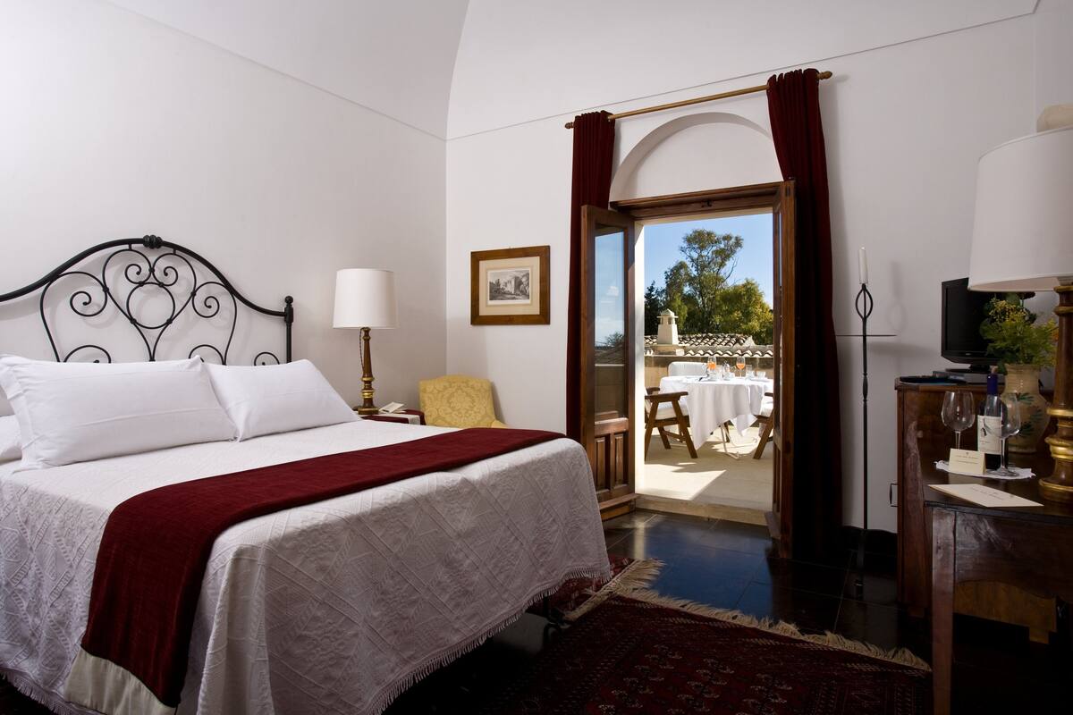 A bedroom with a bed, a bedside table, a lamp, a chair, a TV, and a view of an outdoor patio.