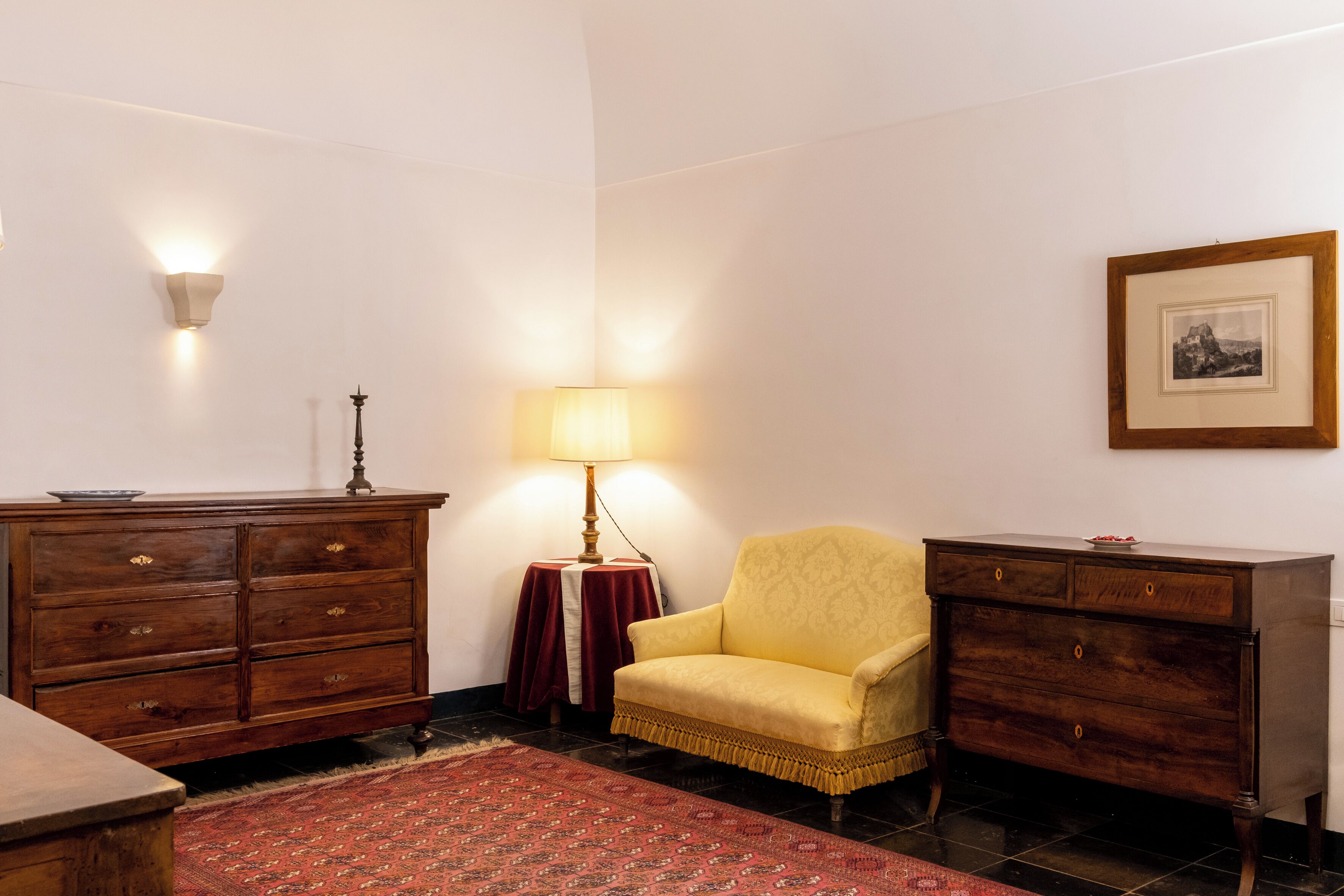 Een kamer met een houten dressoir, een gele fauteuil, een rode vloerkleed, een lamp en een lijst met een afbeelding aan de muur.