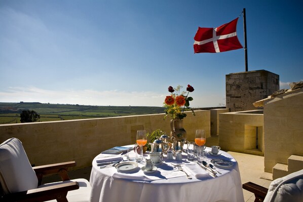 A table set for breakfast with a flag, a vase of flowers, and a view of the countryside.