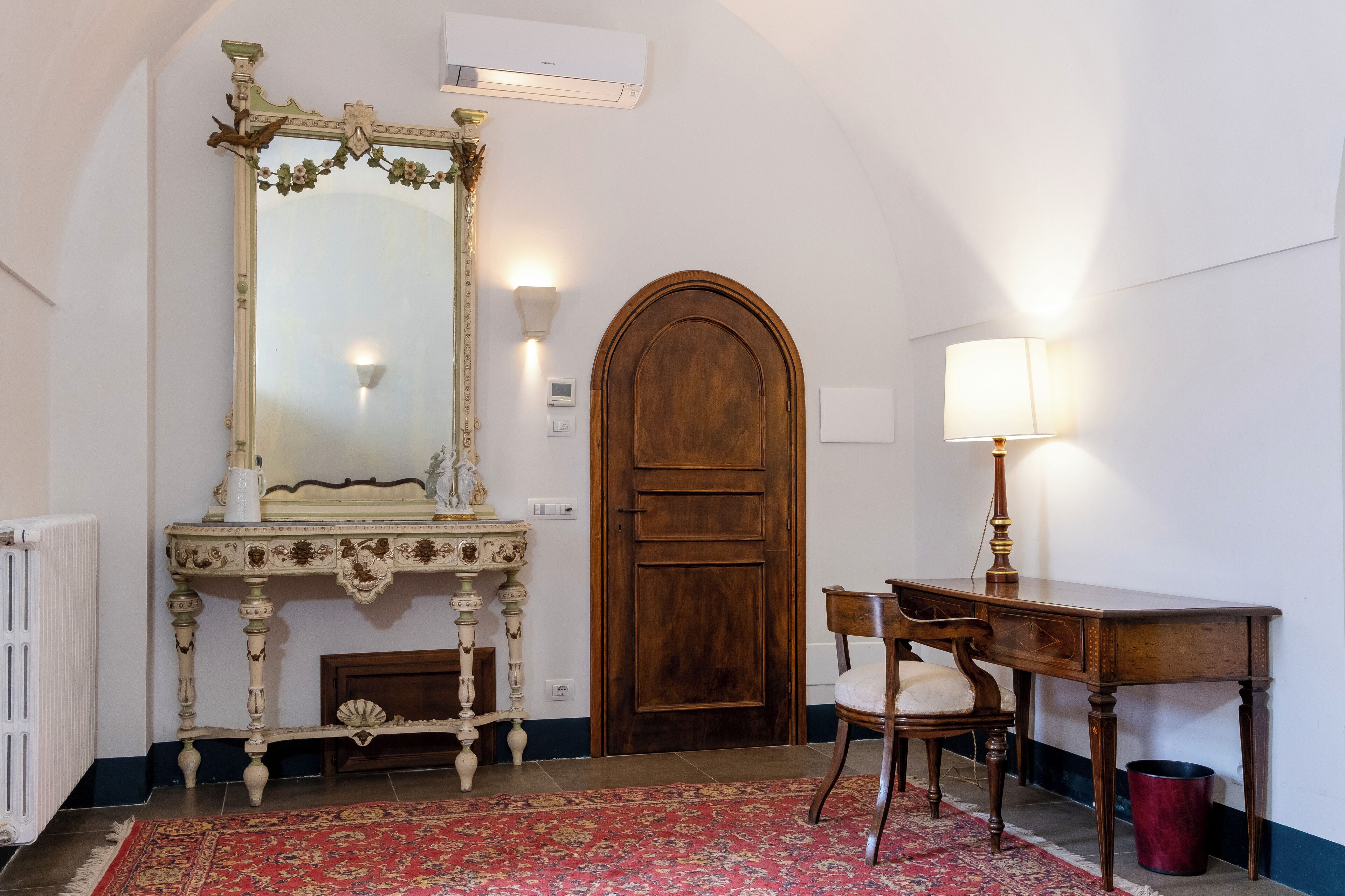 Una habitación con puerta de madera, un espejo grande y decorativo, una consola de madera, una silla y una alfombra roja estampada.