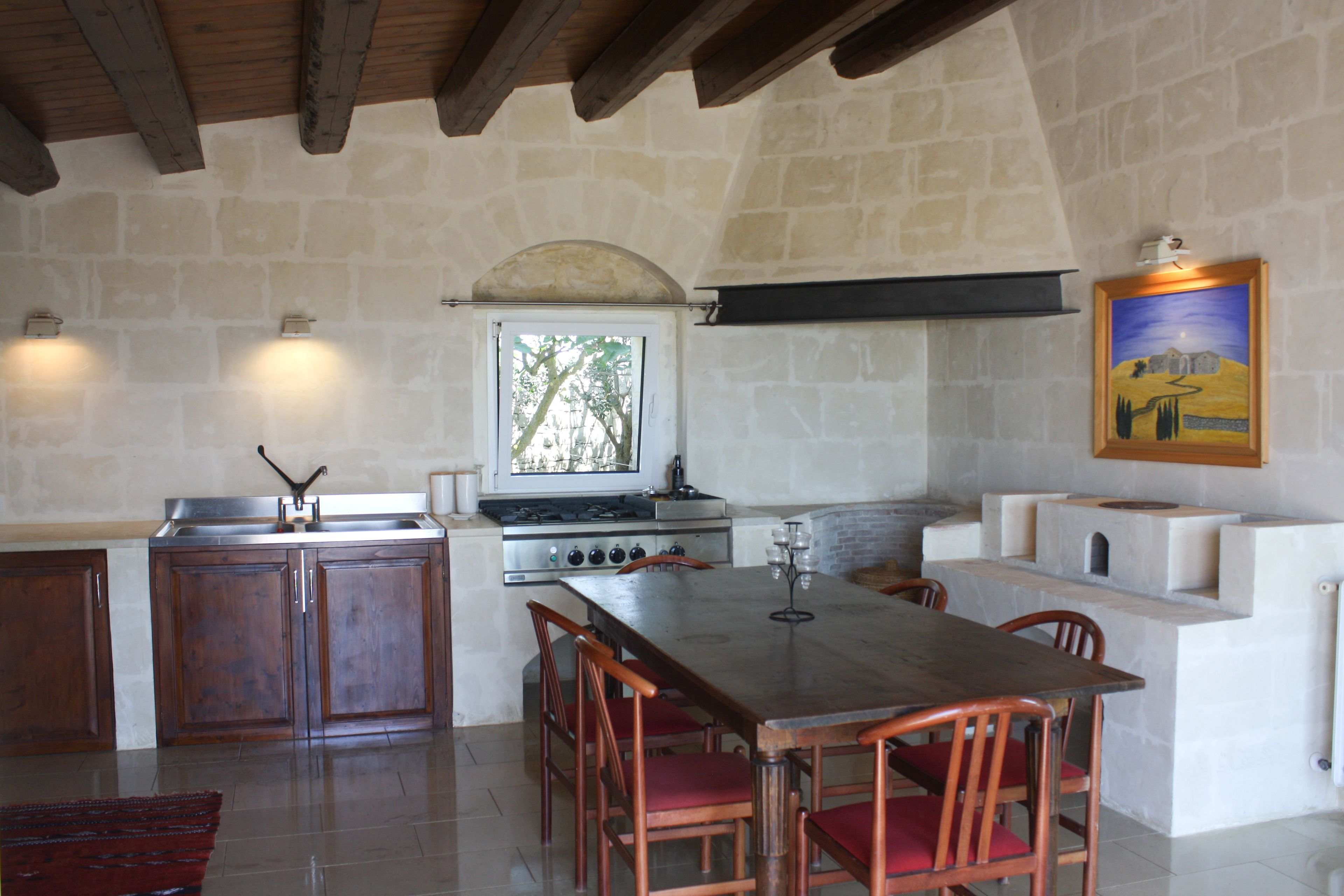 A rustic kitchen with a stone wall, wooden cabinets, a large dining table, and a painting on the wall.