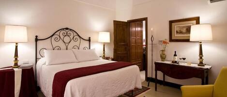 A neatly arranged bedroom with a large bed, two bedside tables with lamps, a small table with a vase and wine glasses, a chair, and a door leading to another room.