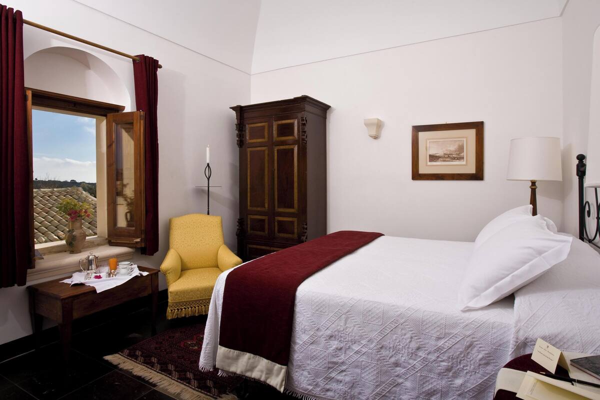 A hotel room with a bed, a chair, a desk with a vase, a cabinet, and a window with curtains.