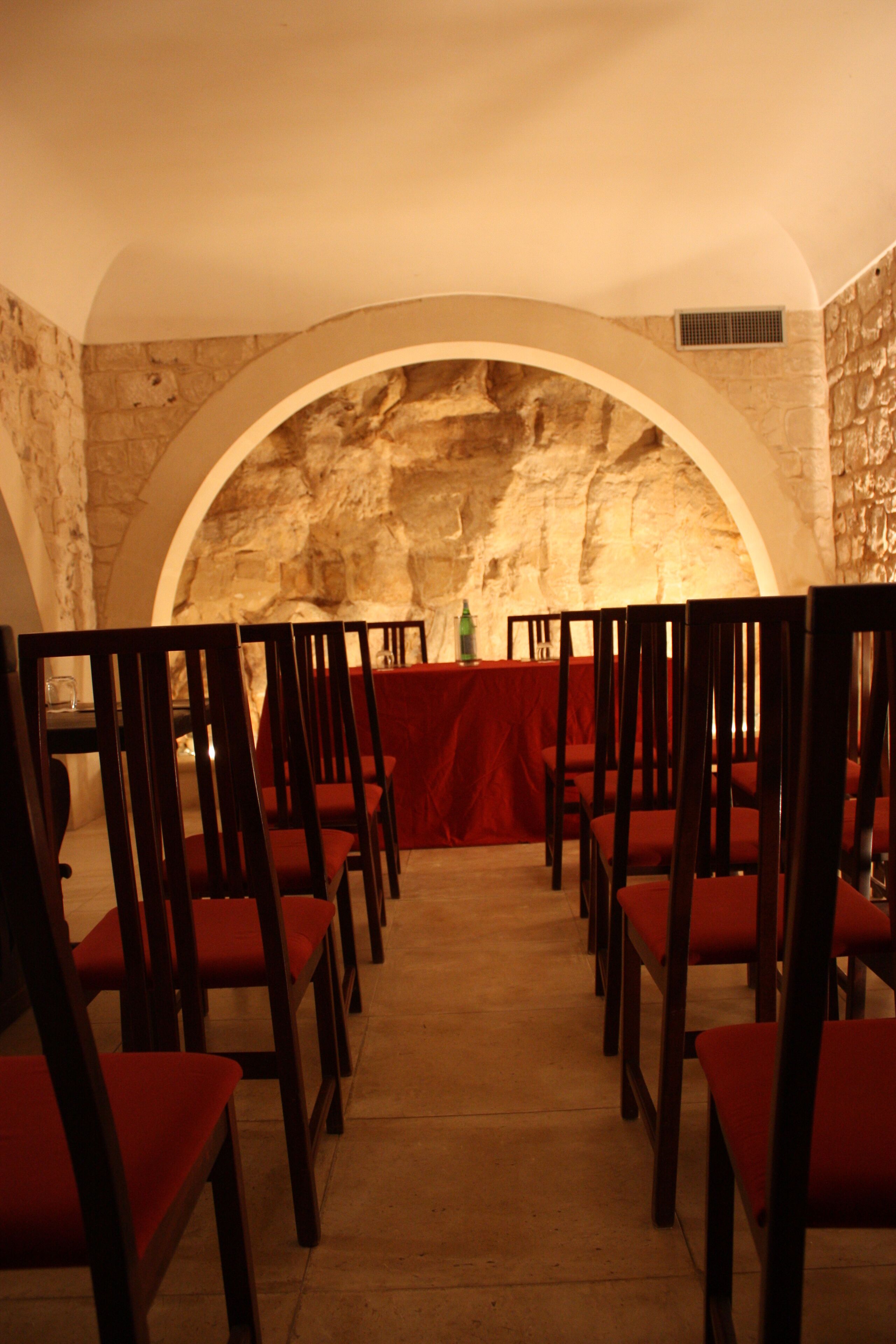 A row of wooden chairs with red cushions arranged in a semi-circular pattern, facing a stone wall with an arched opening.