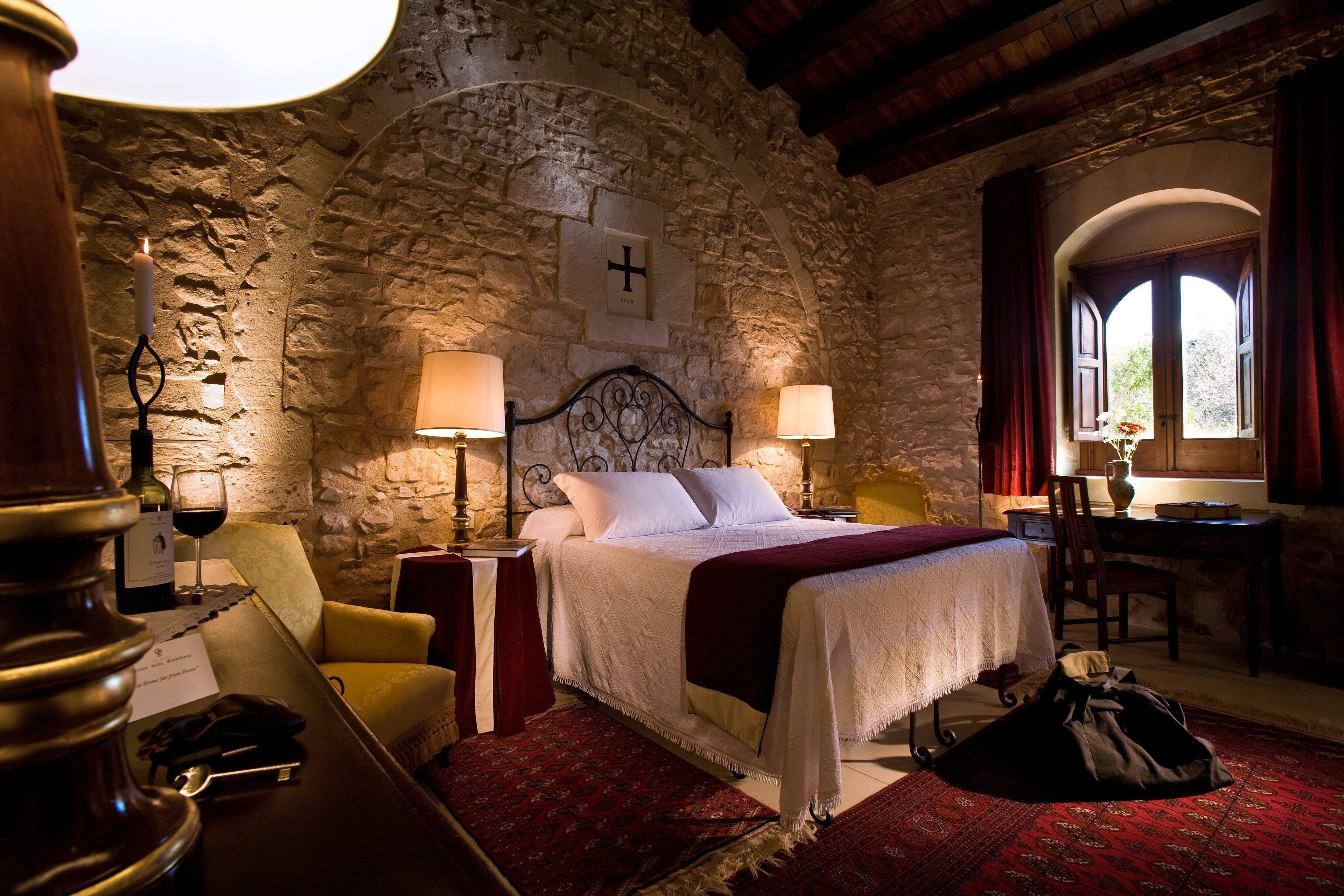 A room with a stone wall, a bed with white linens, two bedside lamps, a wooden chair, a desk with a vase, and a window with curtains.