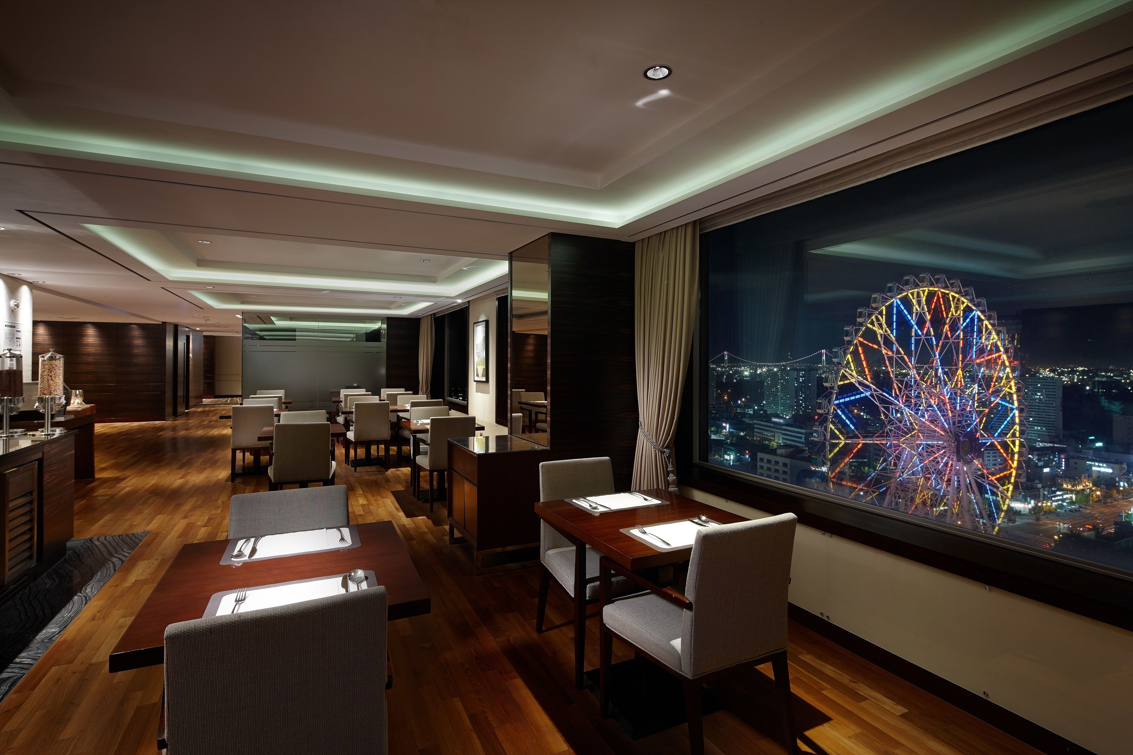 A modern hotel lobby with wooden flooring, dining tables, and chairs, a reception desk, and a large window with a view of a brightly lit Ferris wheel at night.