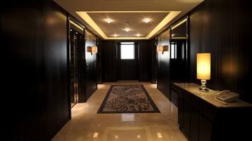 A hallway with a marble floor, dark wood paneling, recessed ceiling lights, a patterned rug, and a desk with a lamp and phone.