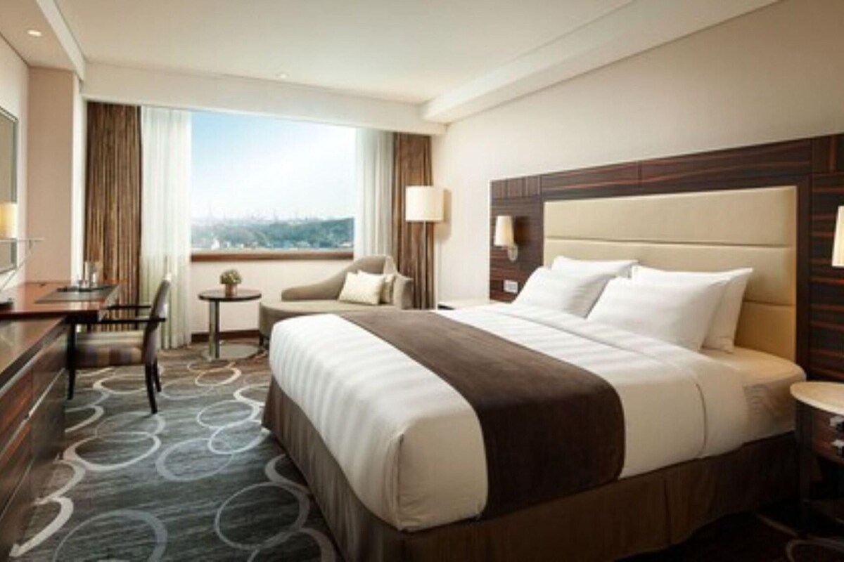 A hotel room with a large bed, a desk, a chair, a small table, and a TV.