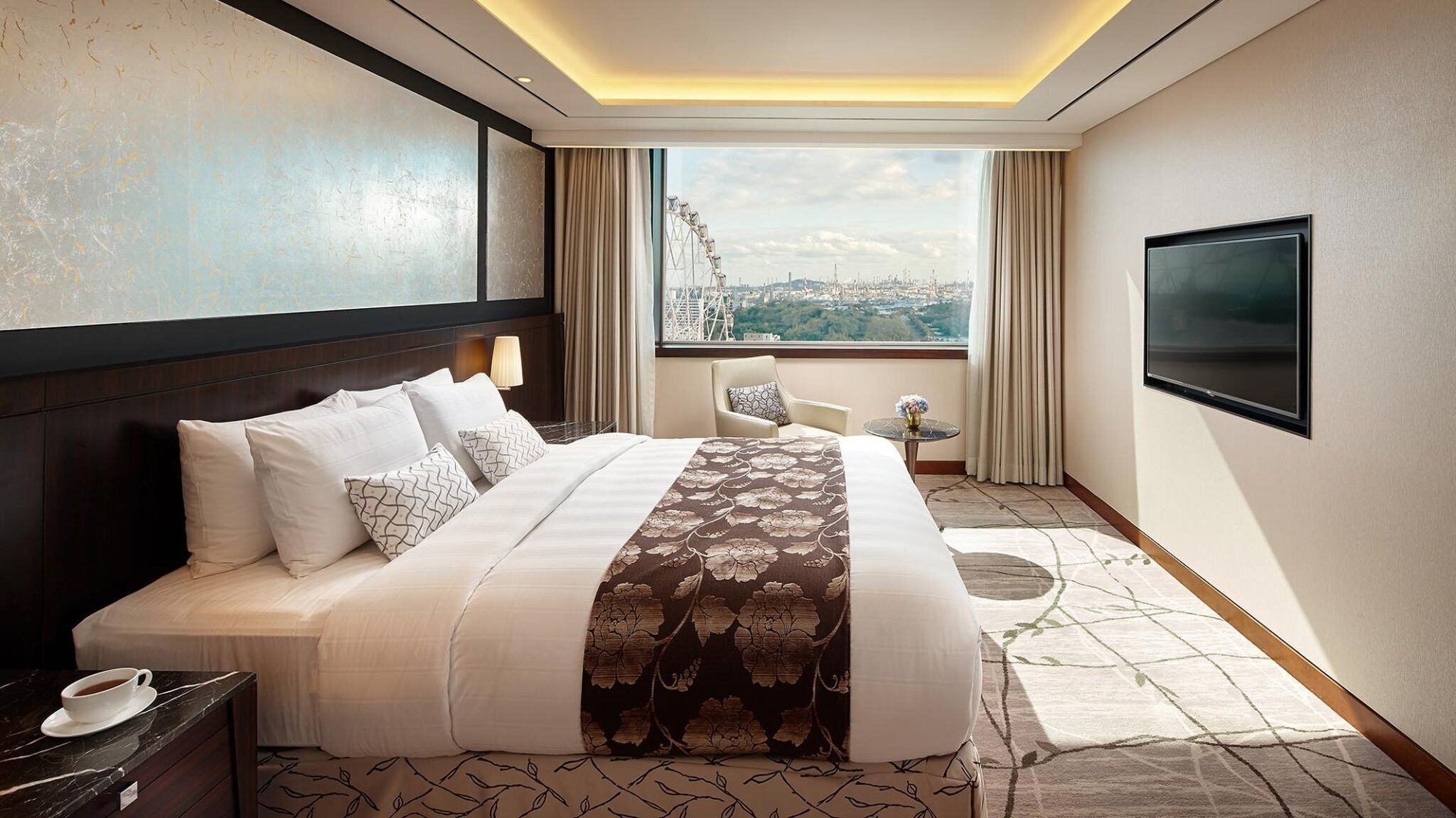 A hotel room with a large bed, a TV, a chair, a table with a vase, and a view of a cityscape.