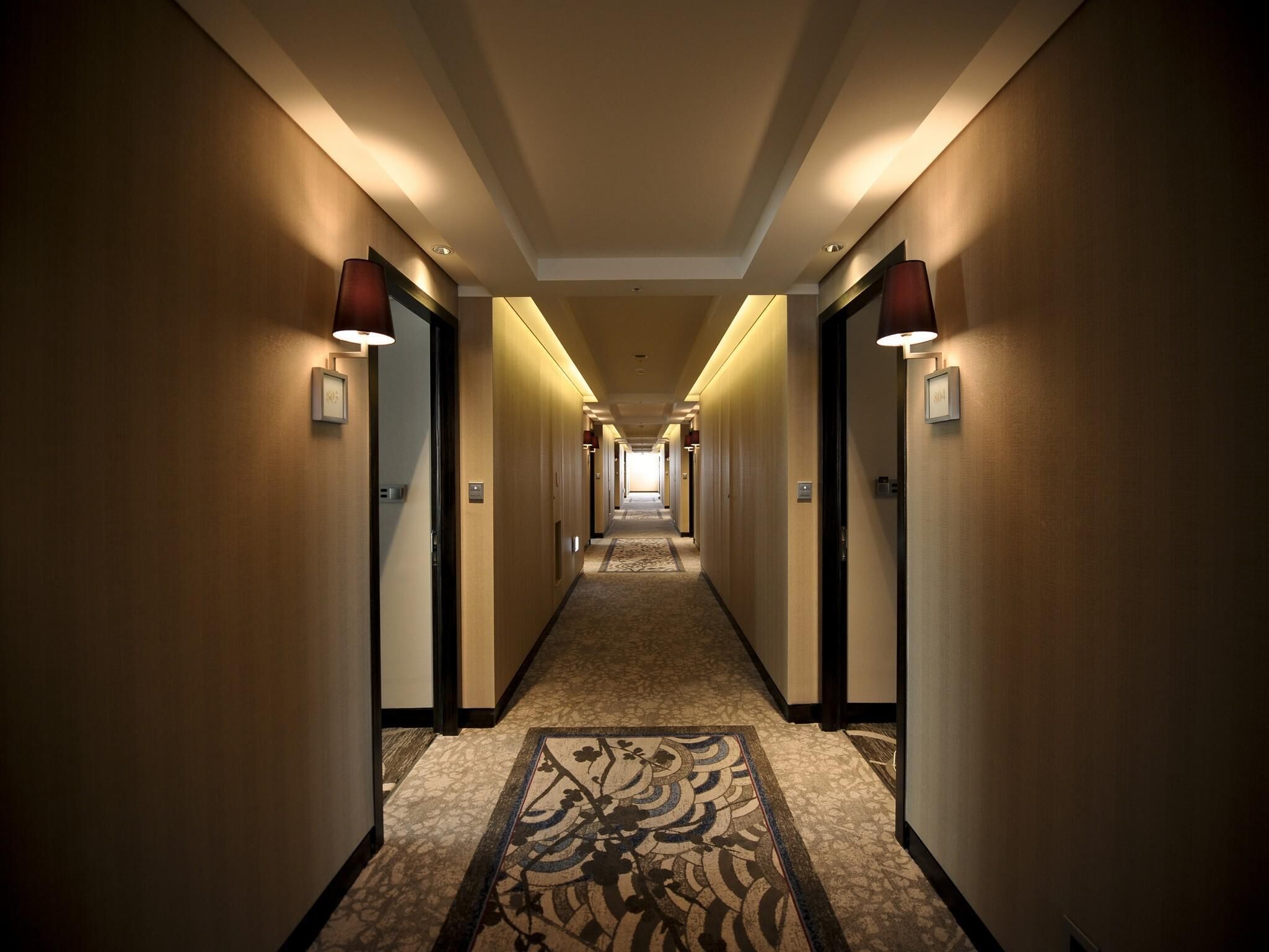 A long, well-lit hallway with doors on both sides, carpeted with a patterned design, and overhead lighting.