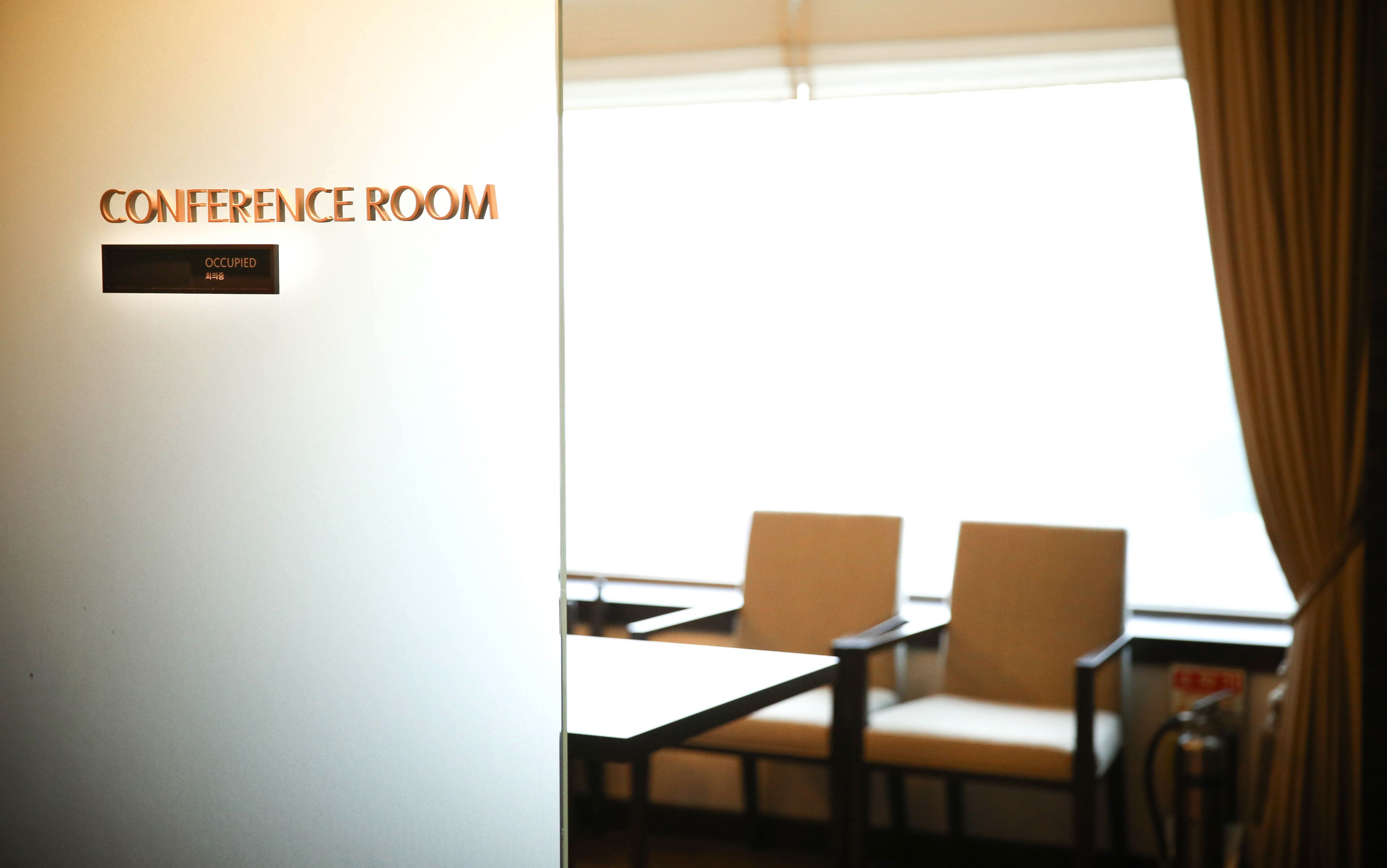 A conference room with two chairs and a table, a sign indicating it is occupied, and a window with curtains.