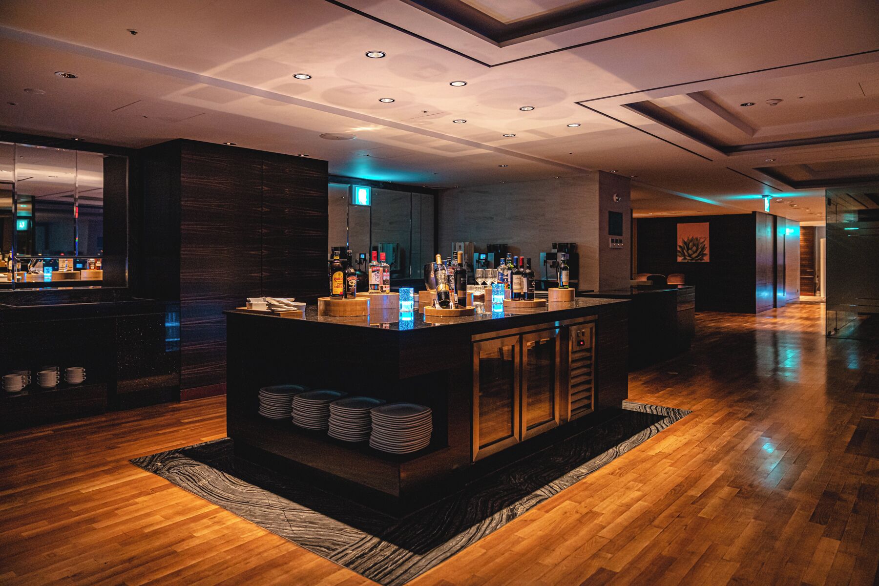 A modern bar area with a wooden counter, stacked plates, and various bottles.