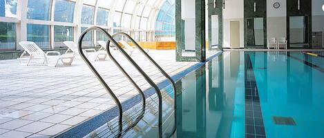 A swimming pool with a glass ceiling, lounge chairs, and a tiled deck area.