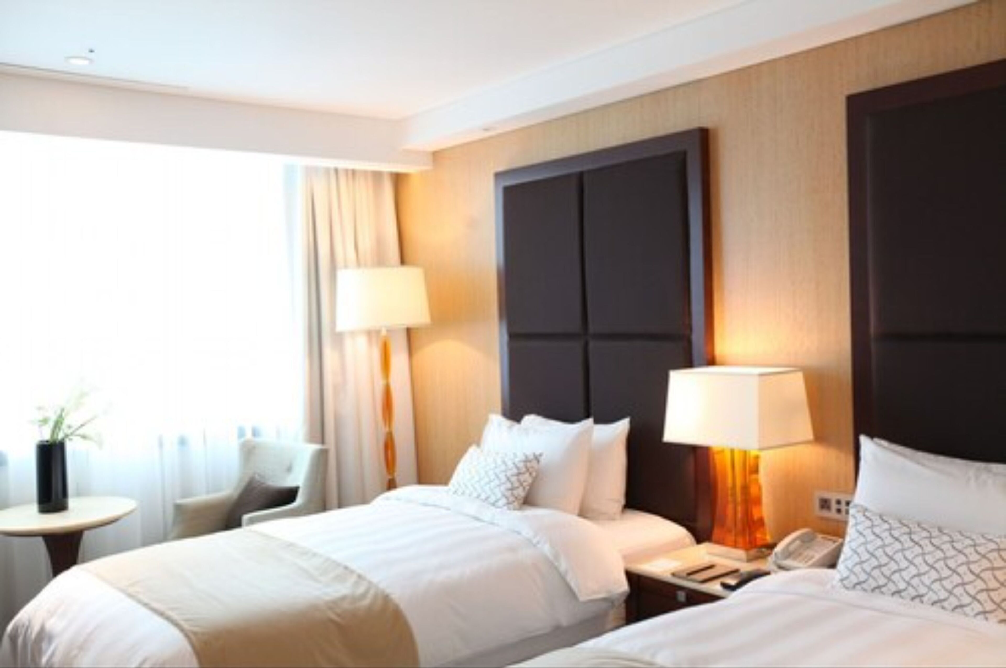 A hotel room with two beds, a nightstand with a lamp, a chair, and a small table with a plant.