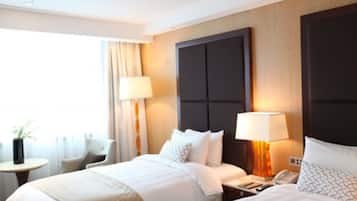 Quarto de hotel com duas camas, mesa de cabeceira com luminária, uma cadeira e uma pequena mesa com uma planta.