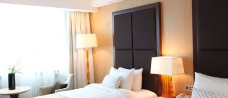 A hotel room with two beds, a nightstand with a lamp, a chair, and a small table with a plant.