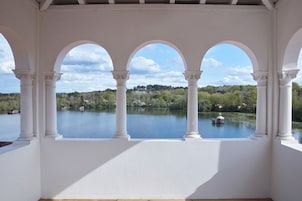A covered balcony with white columns and arches, overlooking a calm lake with a small boat and greenery in the background.