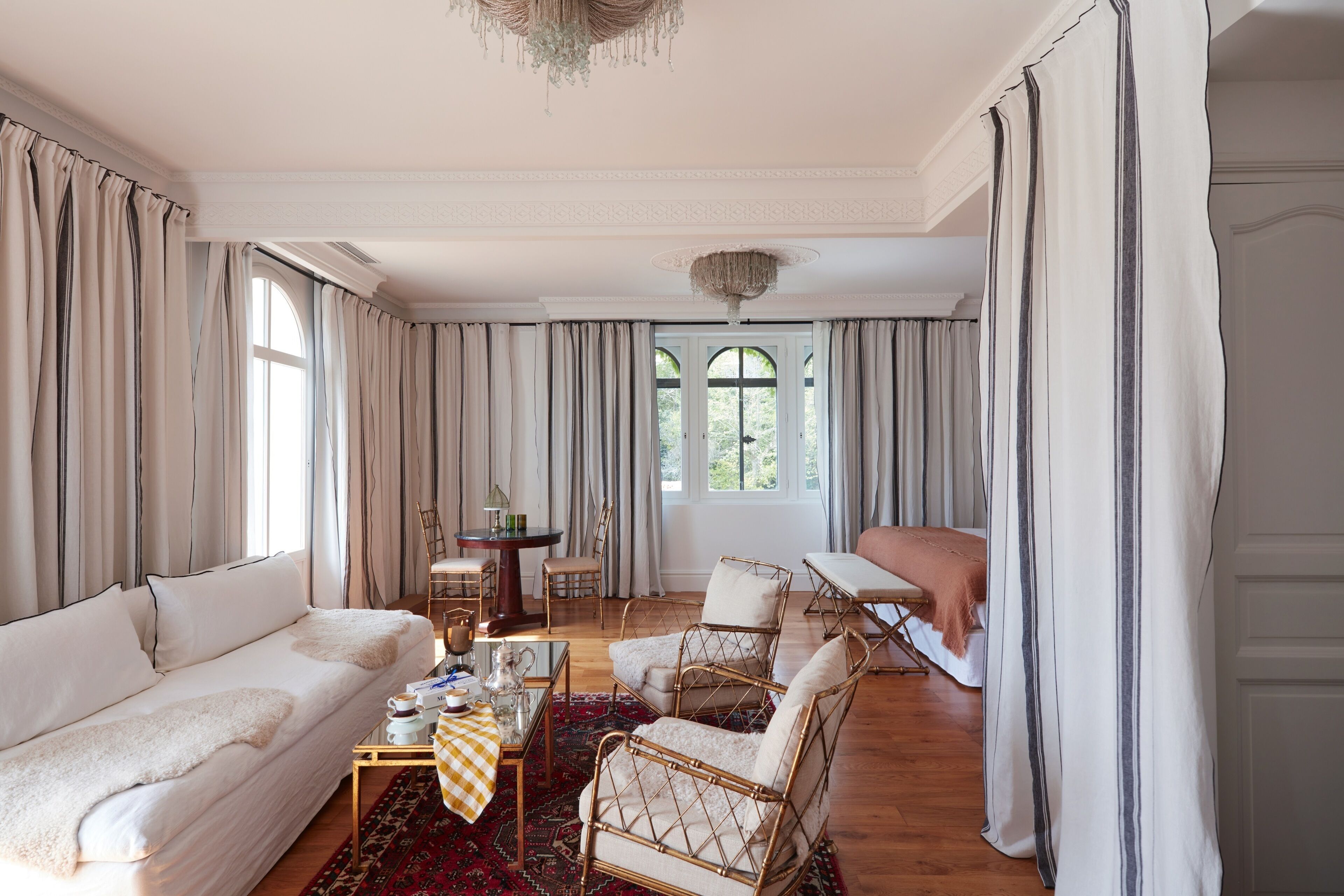 A spacious living room with a sofa, armchairs, a coffee table, and a dining table. There are curtains on the windows, and a chandelier hangs from the ceiling.