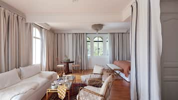 A spacious living room with a sofa, armchairs, a coffee table, and a dining table. There are curtains on the windows, and a chandelier hangs from the ceiling.