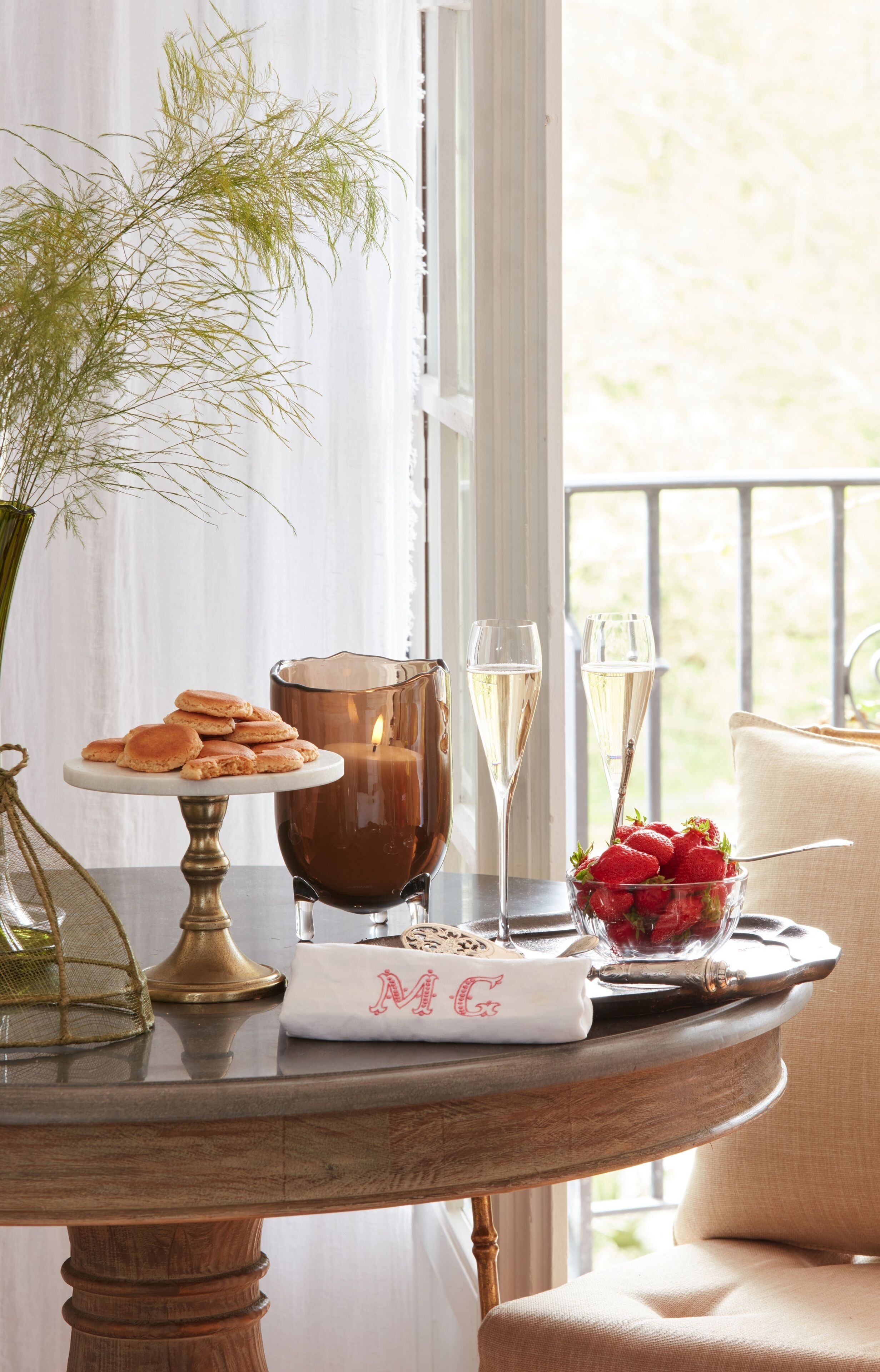 A table with a candle, two glasses of champagne, a plate of pastries, a vase with greenery, and a bowl of strawberries.