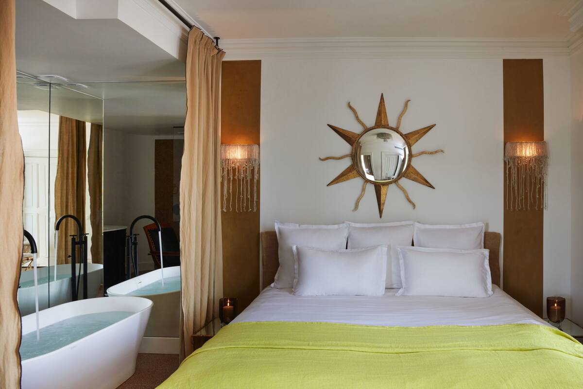 A hotel room with a bed, a large mirror, a sun-shaped wall decoration, and a bathtub.