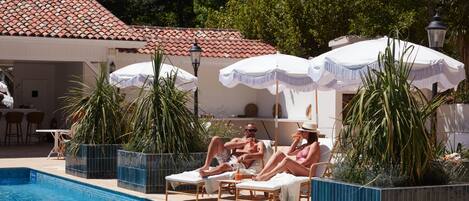 A poolside area with lounge chairs and umbrellas, surrounded by potted plants and a tiled building.
