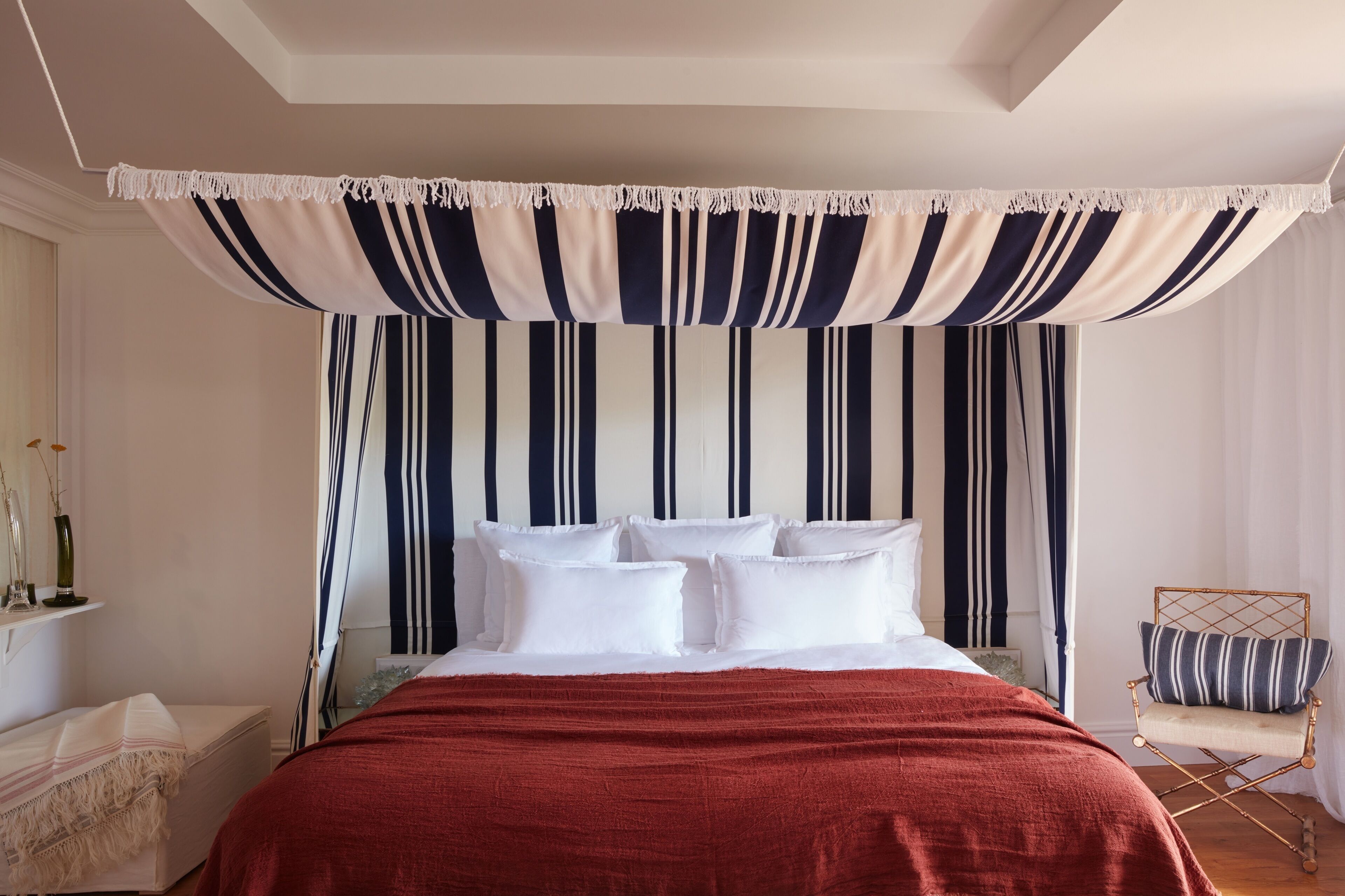 A bedroom with a bed, white pillows, a red blanket, a striped canopy, and a chair with a striped pillow.
