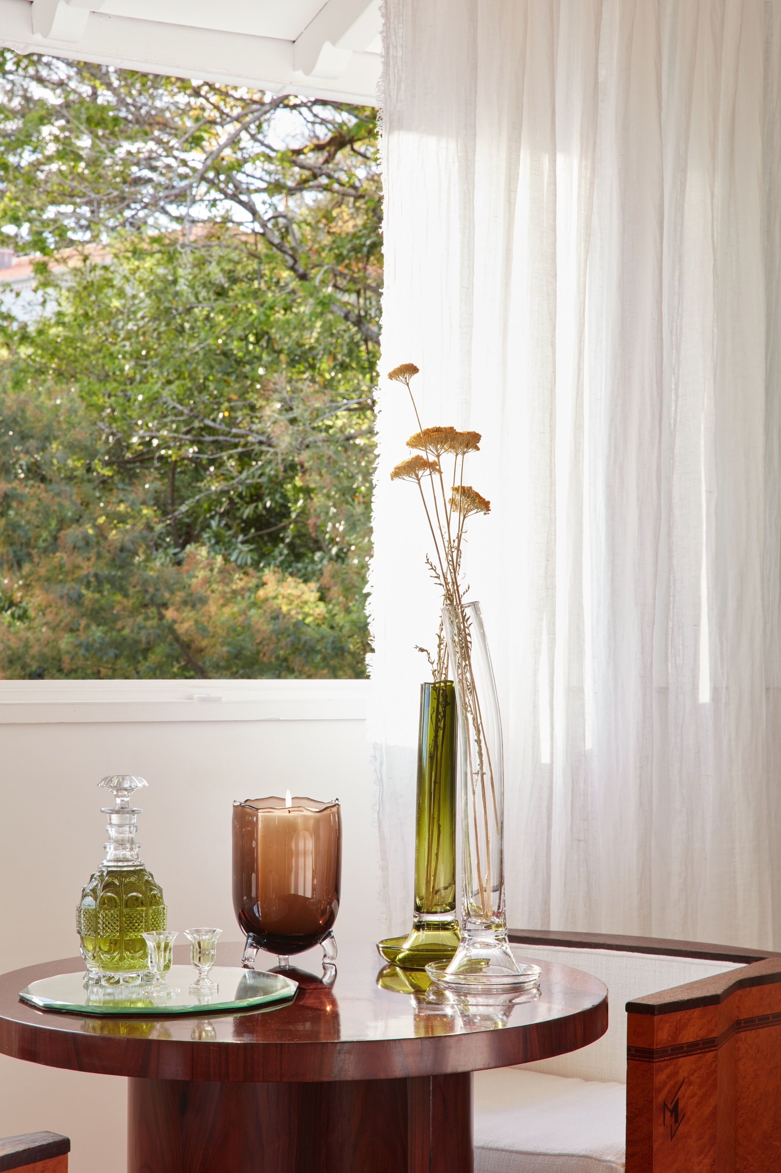 Uma mesa de madeira redonda com um castiçal de vidro, um vaso verde com flores secas e um jarro de vidro.
