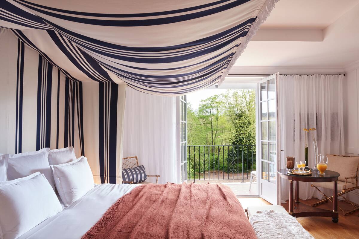 A bedroom with a large bed, a balcony with a view of greenery, and a dining area with a table and chairs.