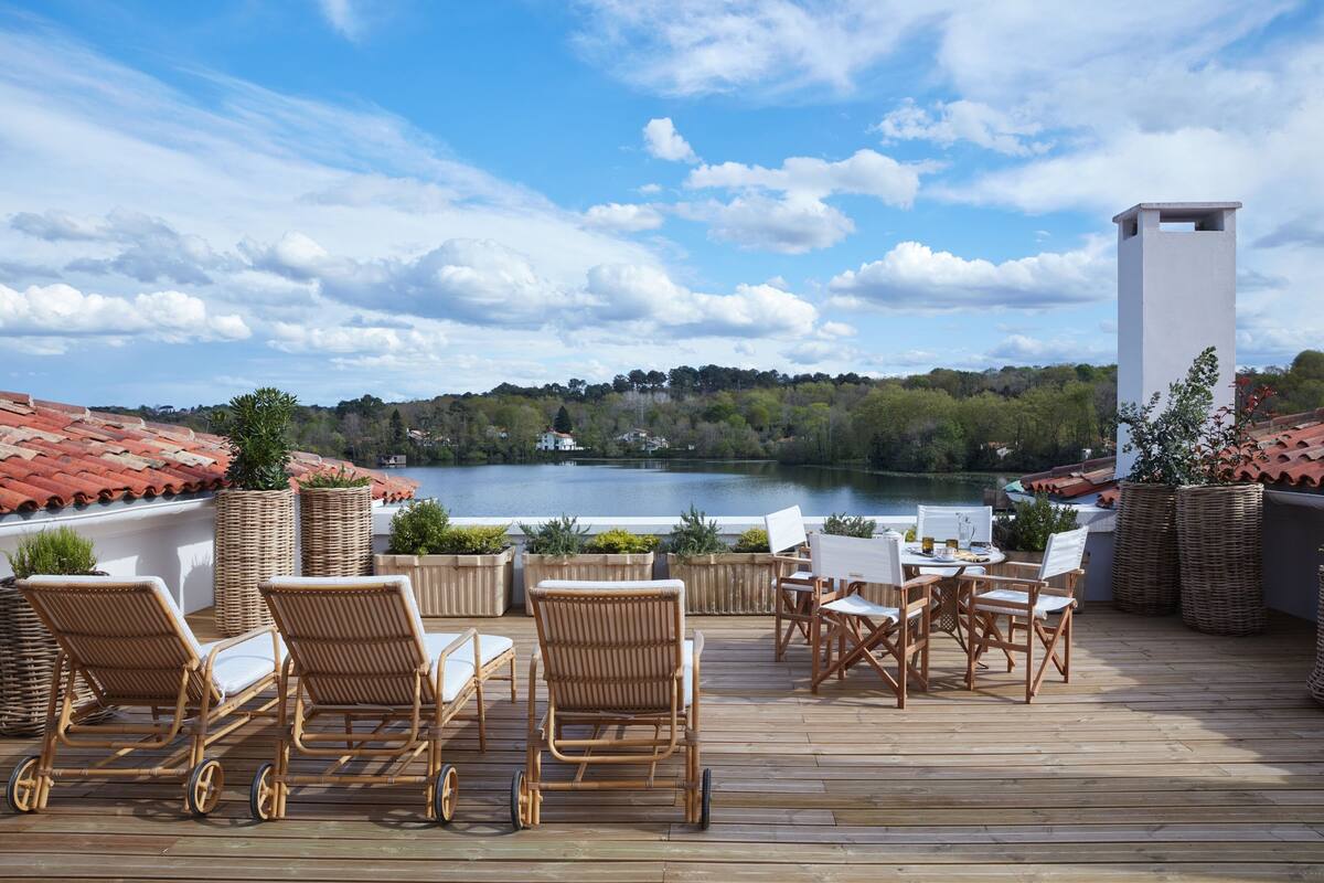 A wooden deck with wicker chairs and a table, overlooking a lake with a red-tiled roof and greenery in the background.
