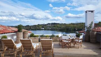 Une terrasse en bois, avec des chaises et une table en osier, donnant sur un lac. On aperçoit, en arrière-plan, un toit recouvert de tuiles rouges et une végétation luxuriante.