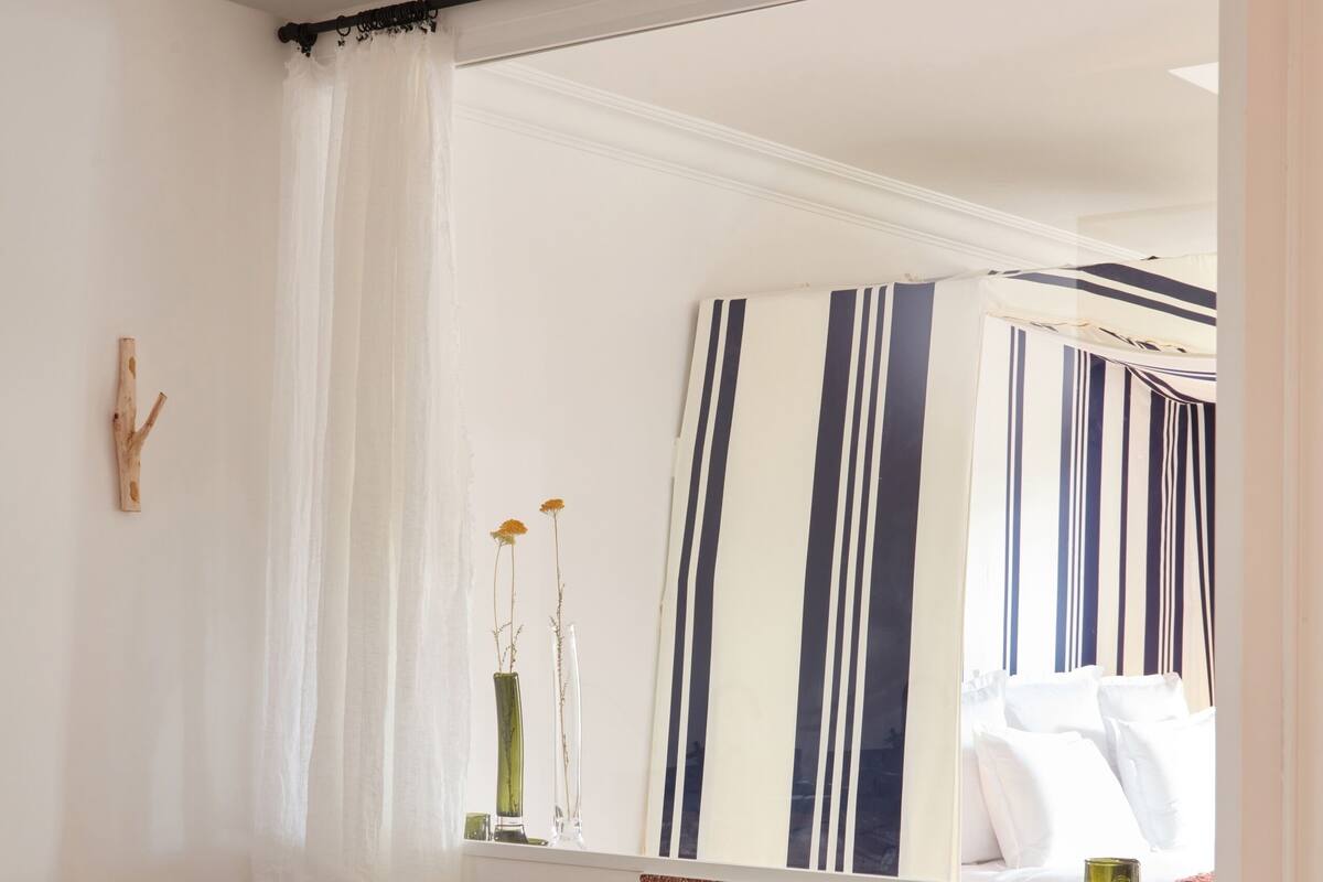 A bathroom with a freestanding bathtub, a wooden stool, a mirror with striped curtains, and a small table with toiletries.