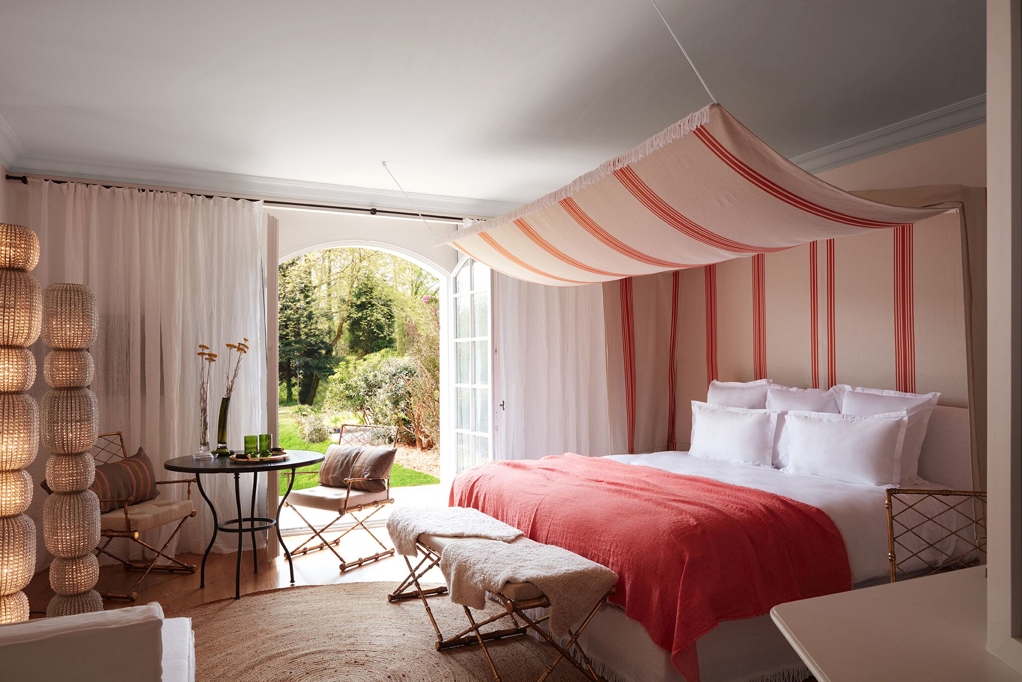 A bedroom with a large bed, a small table, and a chair. There is a window with curtains, a wicker lamp, and a view of greenery outside.