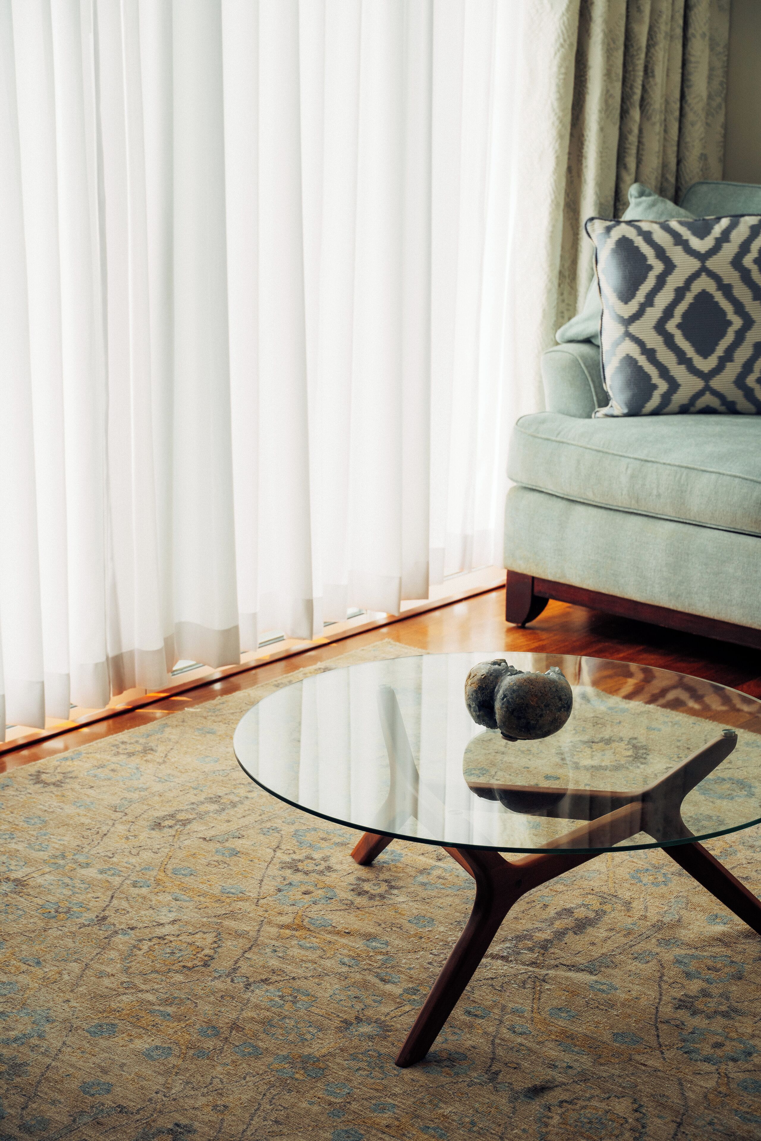 A living room with a glass coffee table, a sofa with patterned cushions, and sheer curtains.