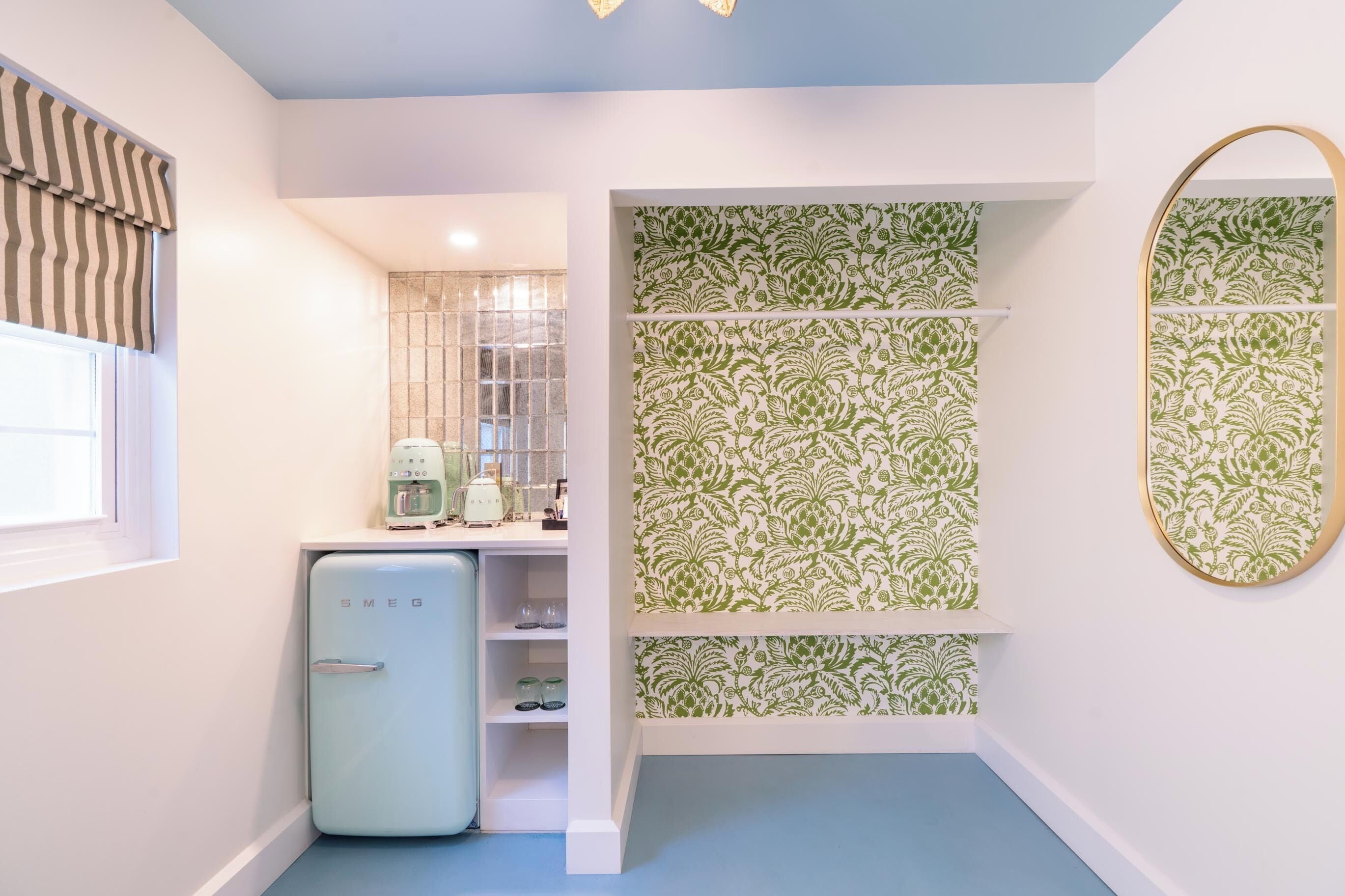 A compact kitchen with a vintage refrigerator, a window with curtains, and a wall-mounted mirror.