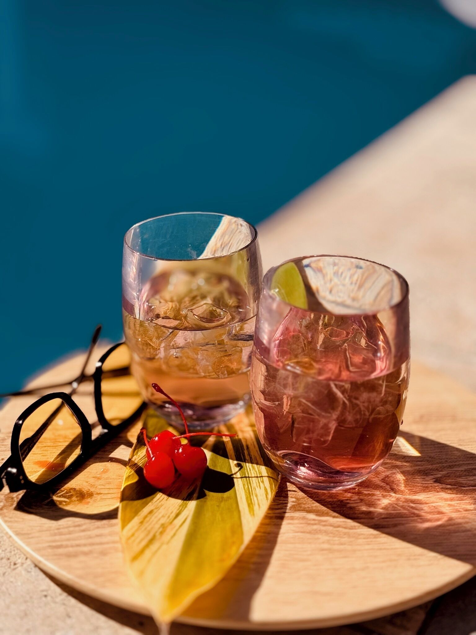Two glasses of beverages on a wooden tray with a pair of glasses and cherries.