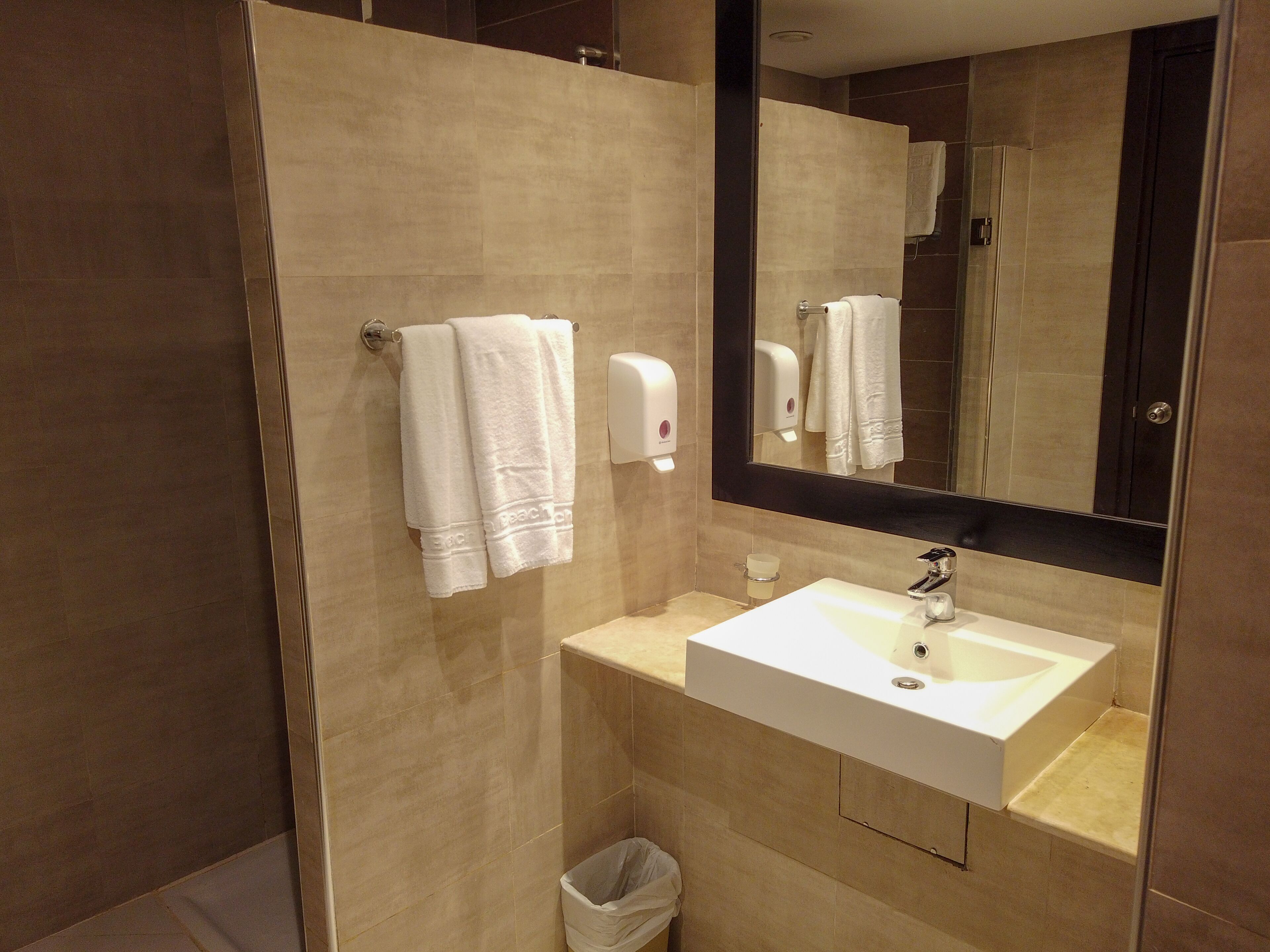 A modern bathroom with a beige tiled wall, a white sink, a mirror, and two towels hanging on a towel rack.