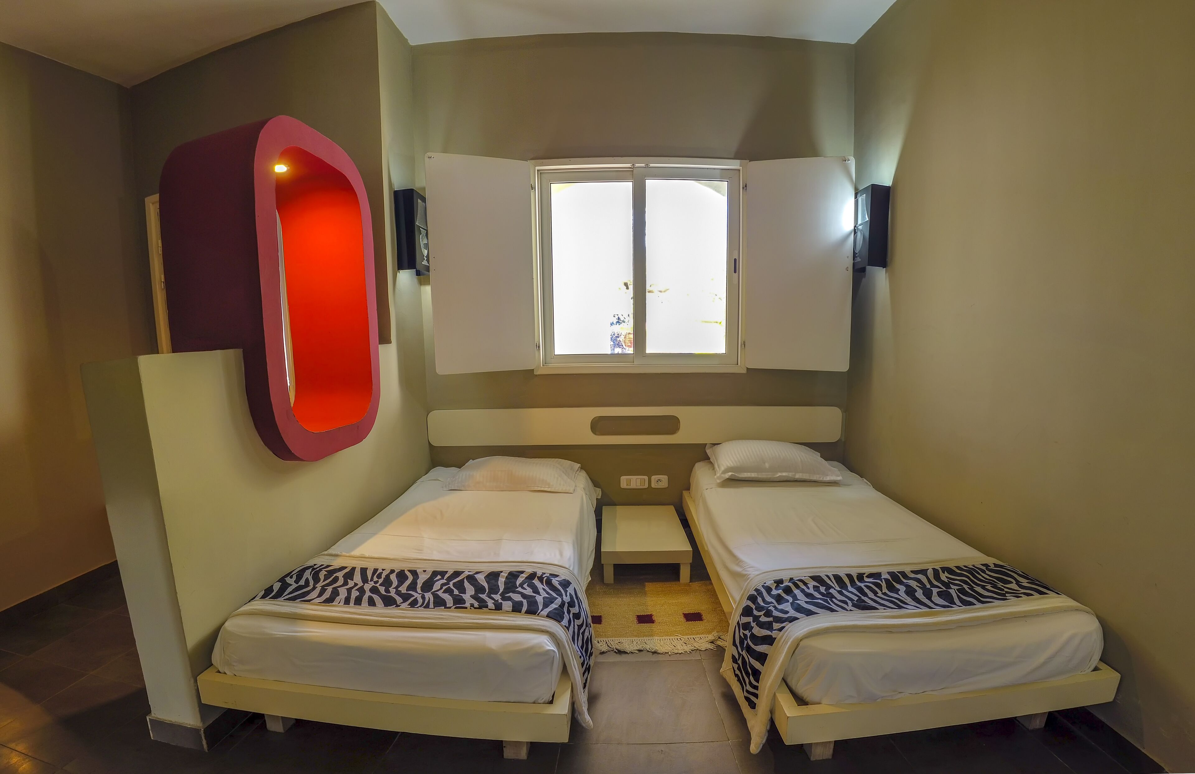 A room with two beds, a window, a red wall feature, and a small table.